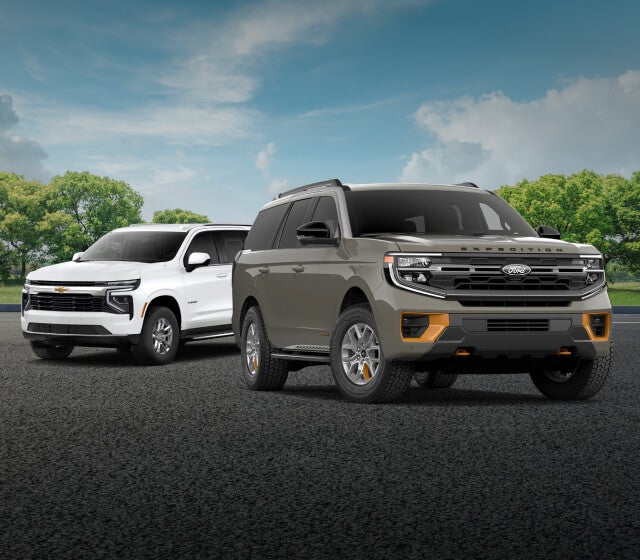 A green Ford Expedition and a white Chevy Tahoe parked in front of a sunny forest background.