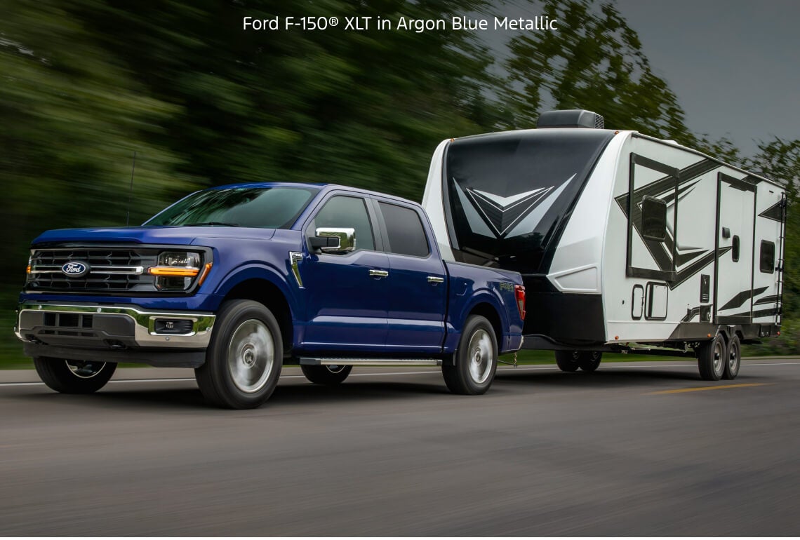 Front quarter tracking view of a Ford F-150 XLT in Argon Blue Metallic towing a large travel trailer on a highway.