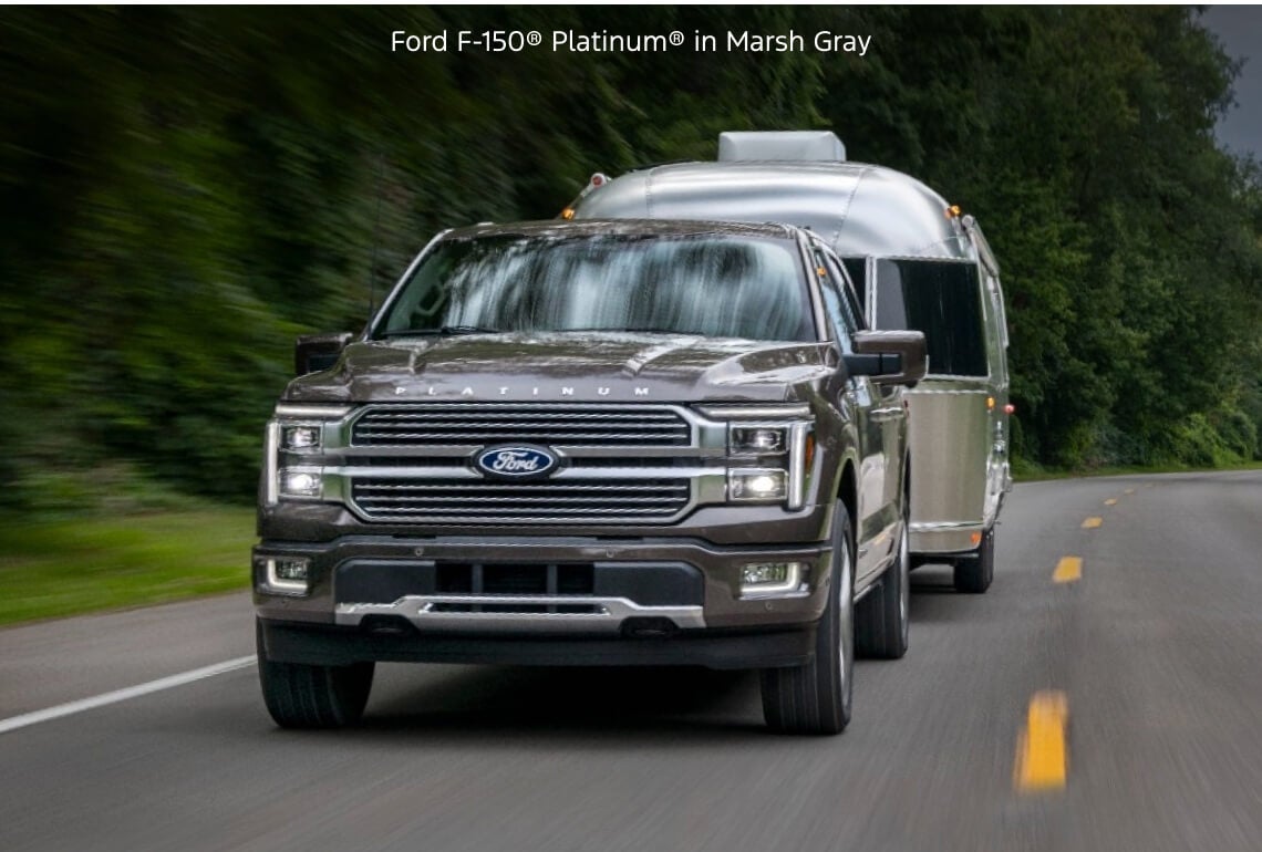 Front tracking view of a Ford F-150 Platinum in Marsh Gray towing an Airstream trailer down a wooded highway.