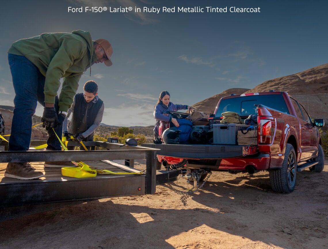 Rear view of a Ford F-150 Lariat in Ruby Red Metallic with its tailgate open while people unload outdoor gear in the desert.